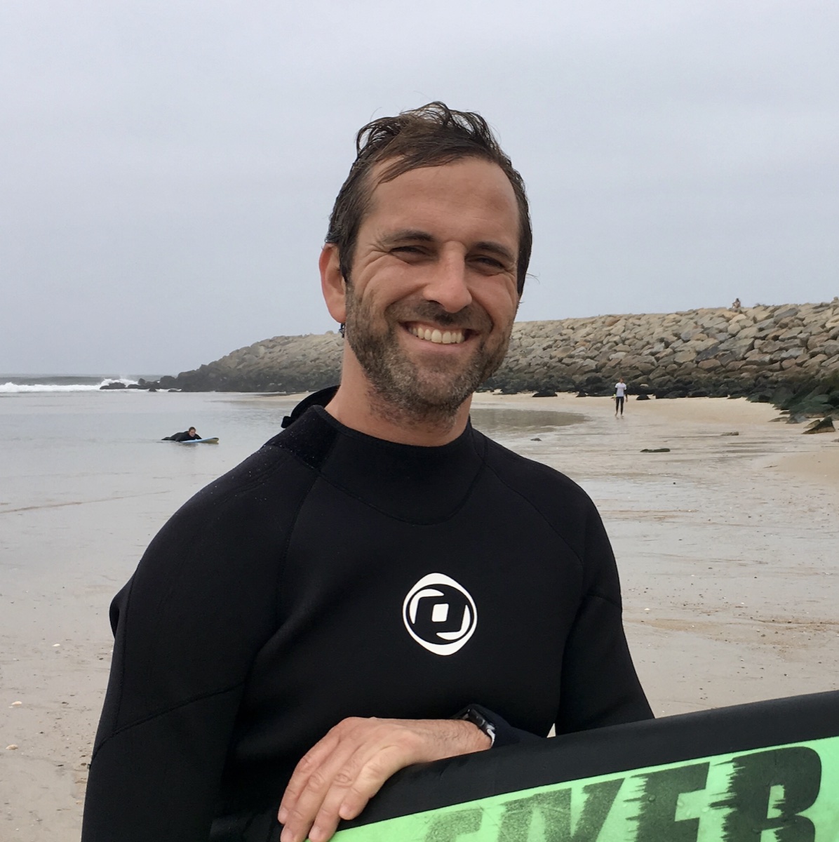 Pedro Cardoso smiling on a beach in a wetsuit beside a surfboard